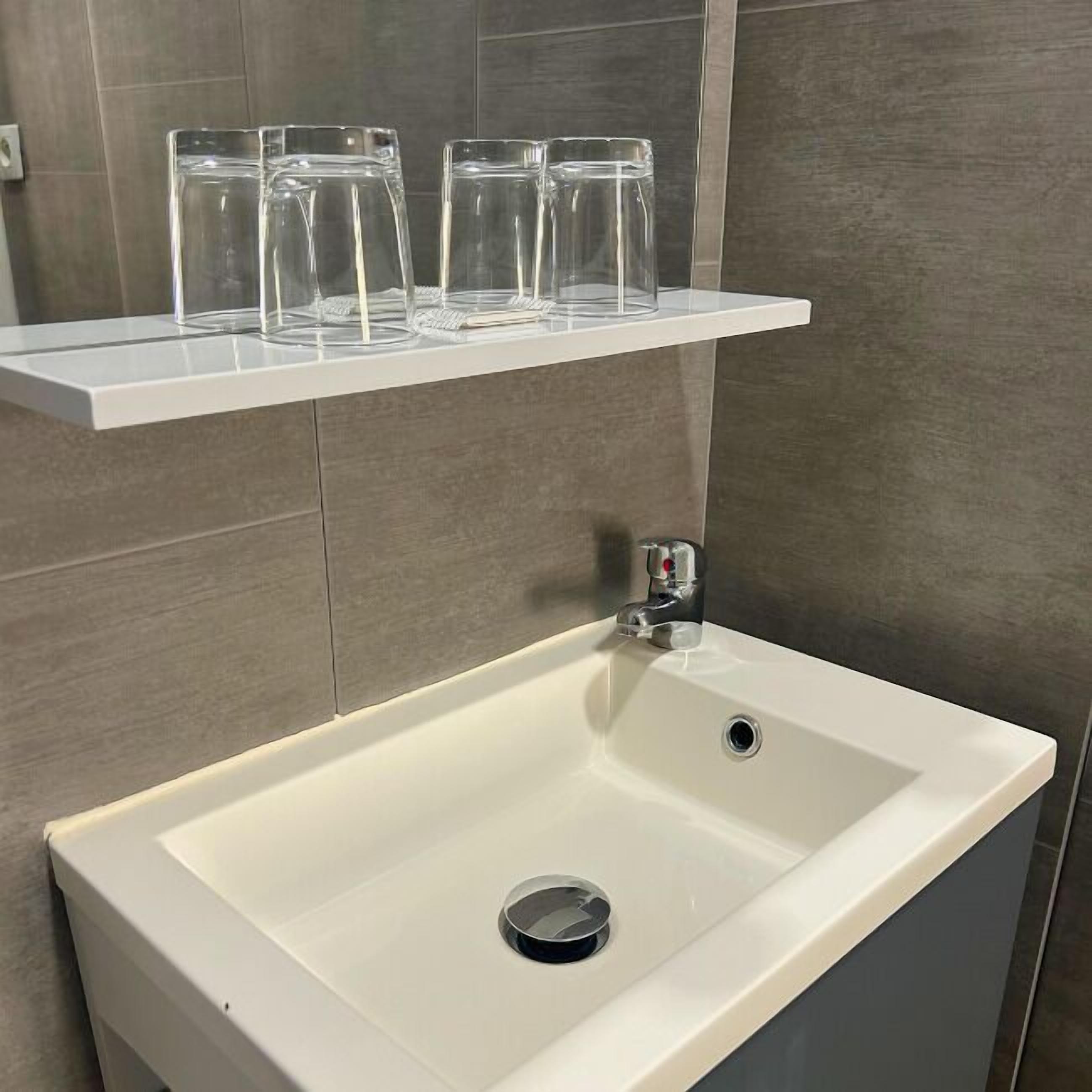 Une salle de bain avec un lavabo blanc, un miroir et une étagère au-dessus du lavabo où sont posés trois verres transparents.