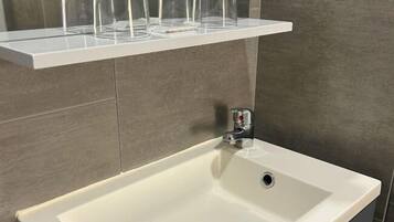 Une salle de bain avec un lavabo blanc, un miroir et une étagère au-dessus du lavabo où sont posés trois verres transparents.
