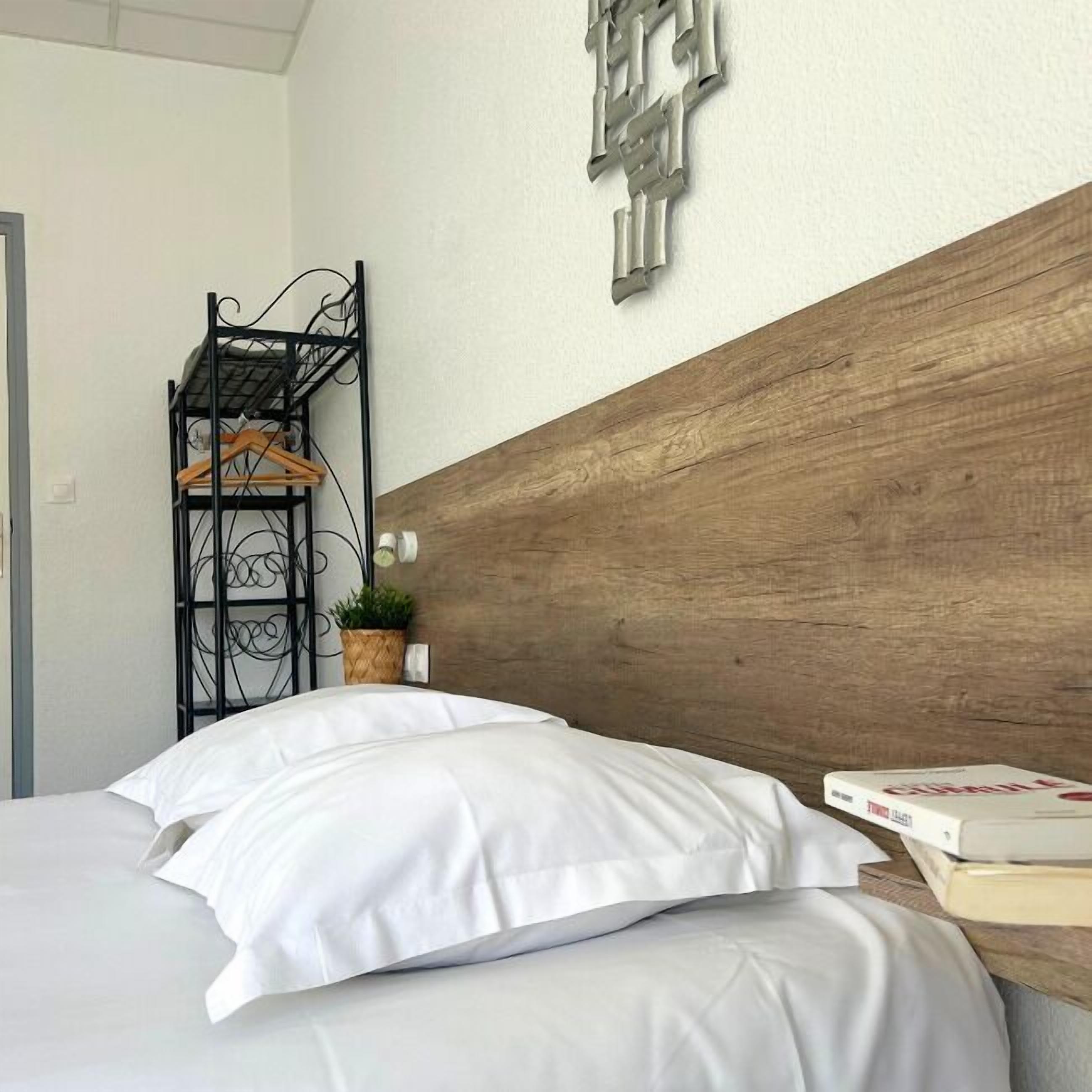 A neatly made bed with white bedding, a wooden headboard, and a small bedside table with a book and a potted plant.