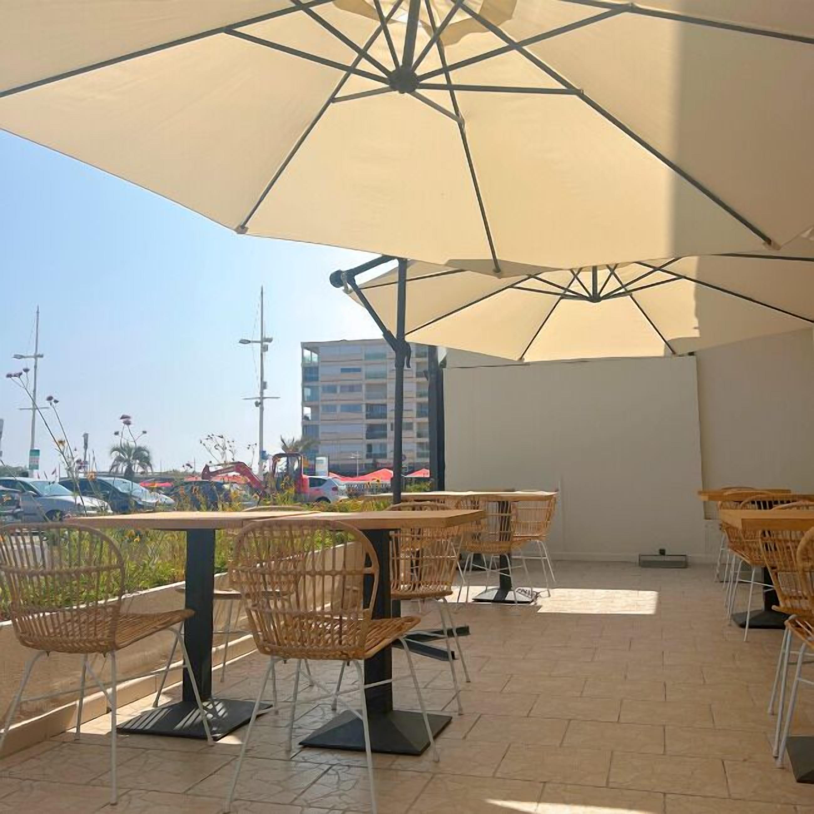 An outdoor patio area with multiple tables and chairs, a large umbrella providing shade, and a view of the surrounding area including parked cars and buildings.