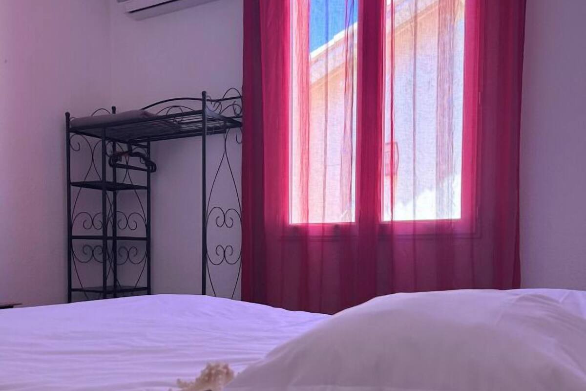 A room with a bed, a metal bunk bed, a window with pink curtains, and an air conditioning unit on the wall.
