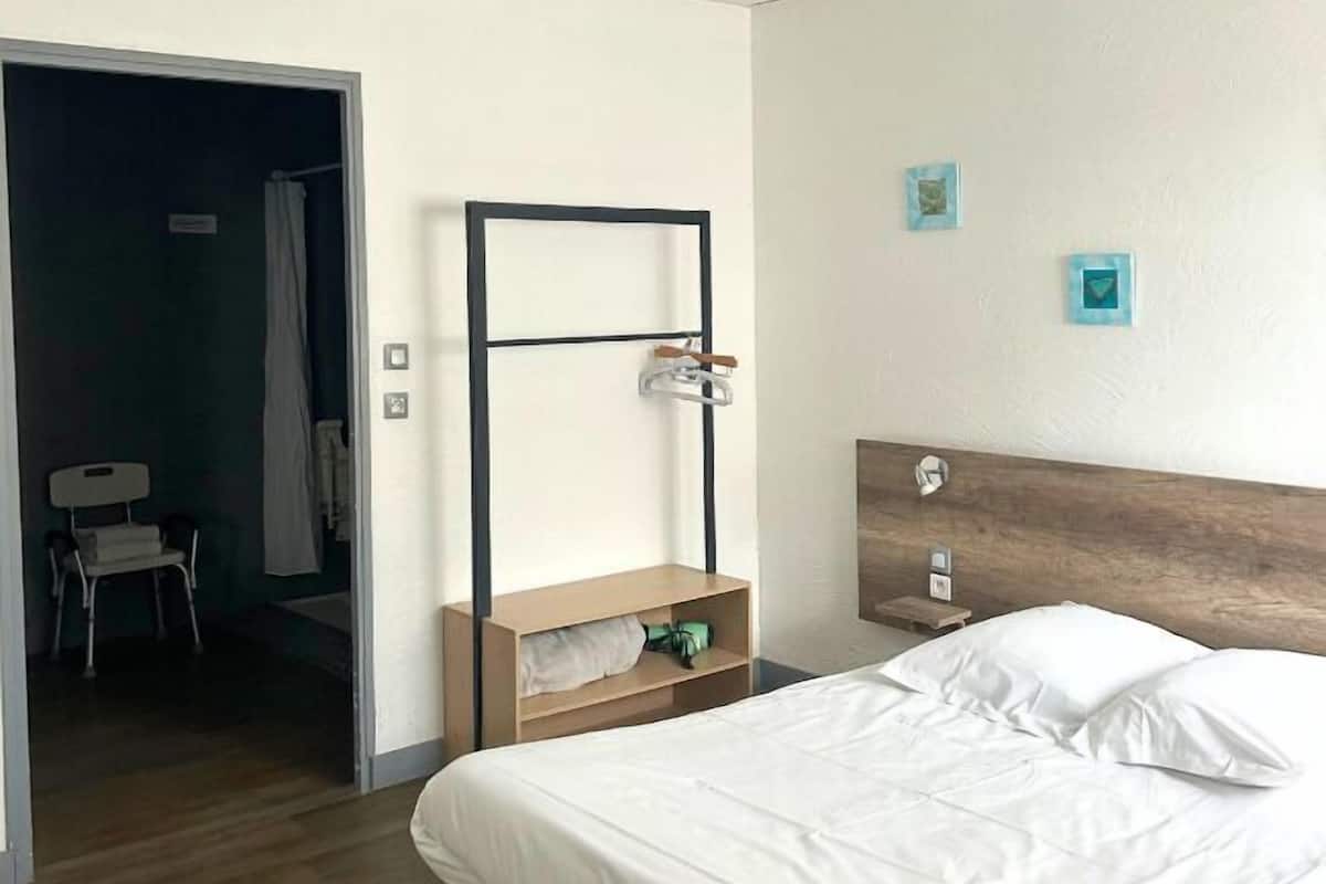 A hotel room with a bed, a wooden headboard, a bedside table with a lamp, and a small shelf. There is a chair in the corner and a door leading to another room.