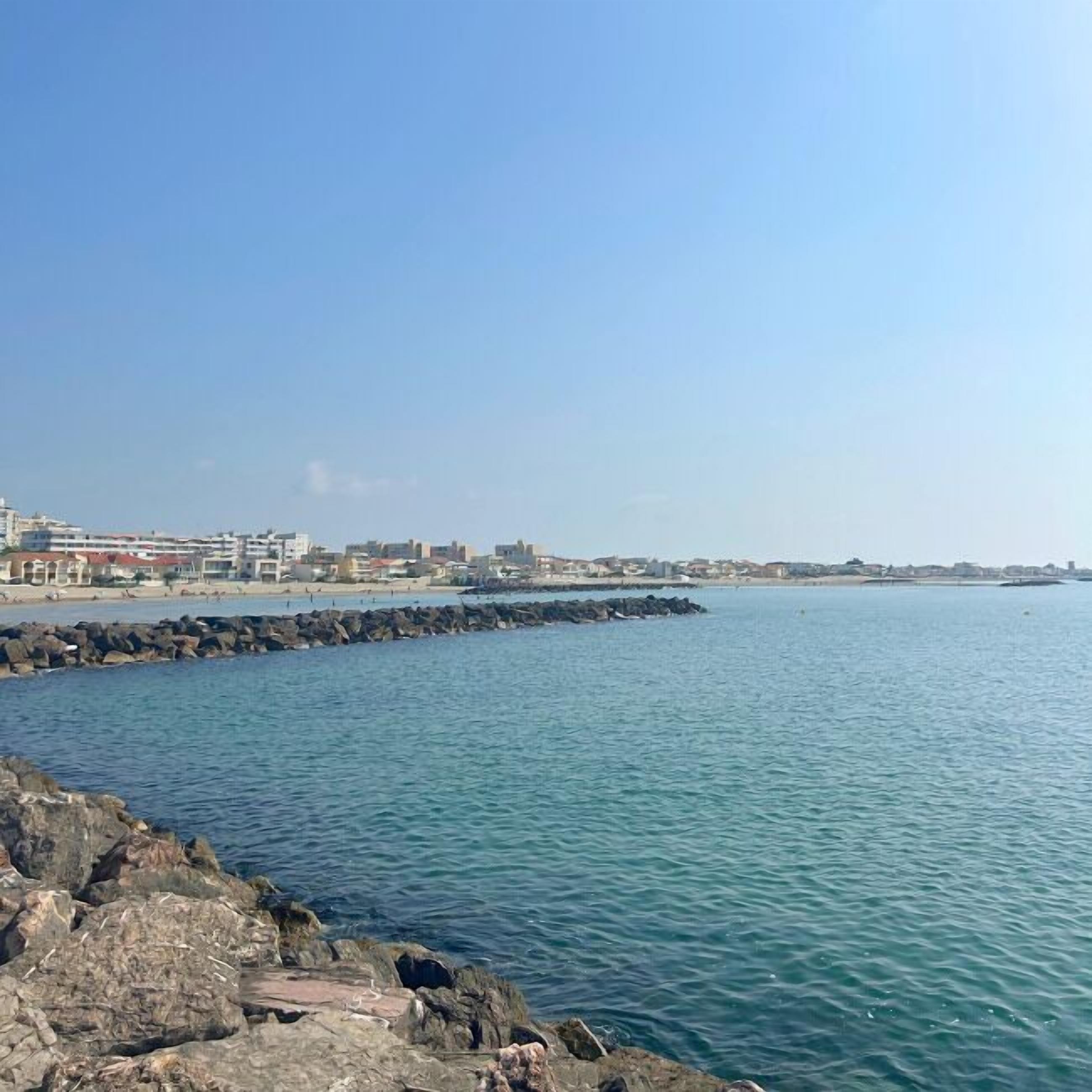Vista sulla costa con un molo roccioso, mare calmo e edifici sullo sfondo.