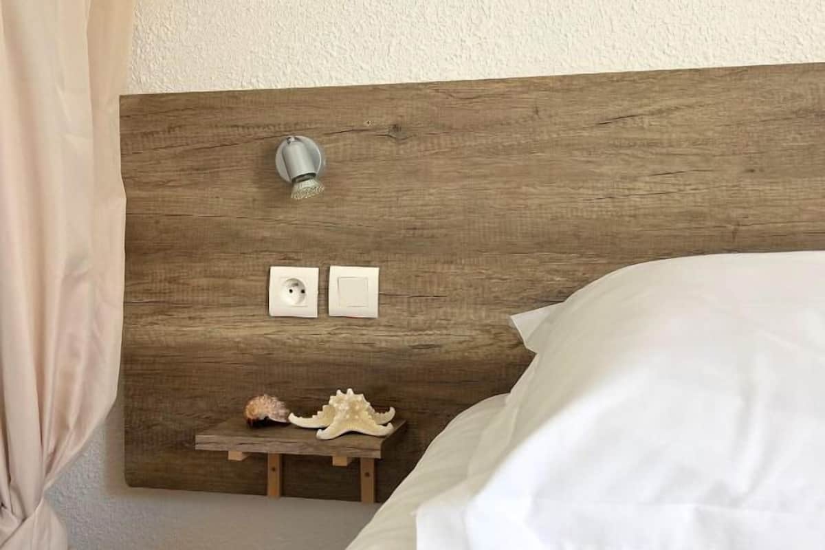 A bedroom with a wooden headboard, a bedside lamp, two wall sockets, and decorative shells on a small wooden shelf.