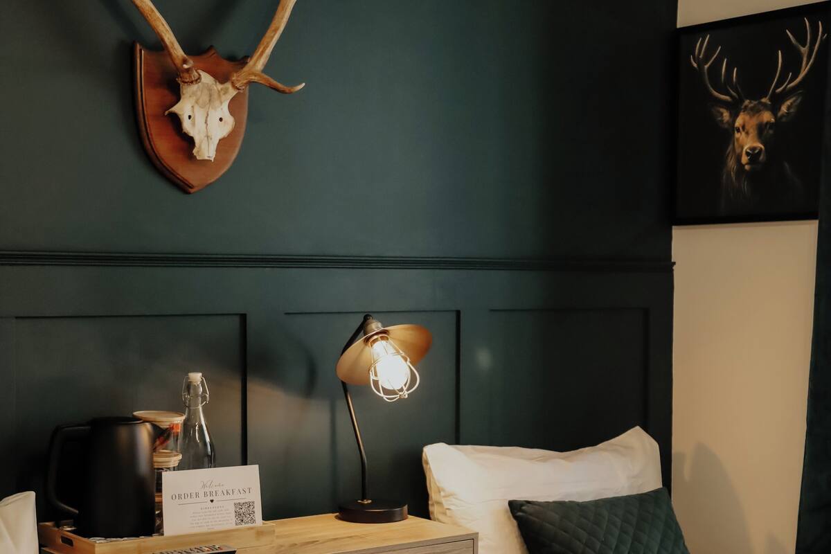 A hotel room with a bed, a wooden dresser, a wall-mounted deer skull, a lamp, and a sign indicating breakfast service.