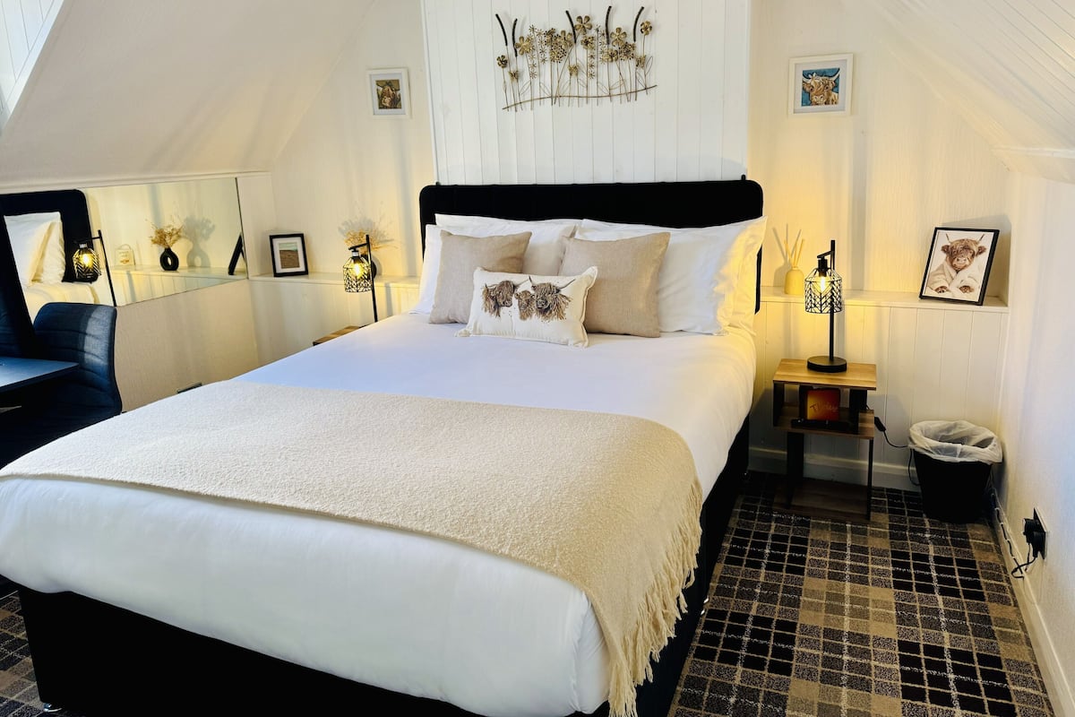 A neatly arranged bedroom with a large bed, a bedside table with a lamp, and a small shelf with pictures.