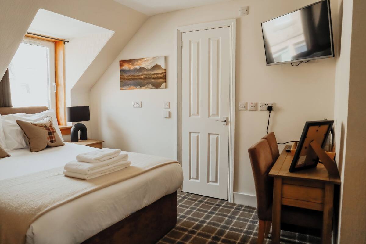 A bedroom with a bed, a desk, a chair, a TV, a door, and a painting on the wall.