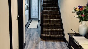 Un couloir avec un plancher en bois, un escalier recouvert d’une moquette à carreaux, un vase rempli de fleurs et un bureau avec un livre.