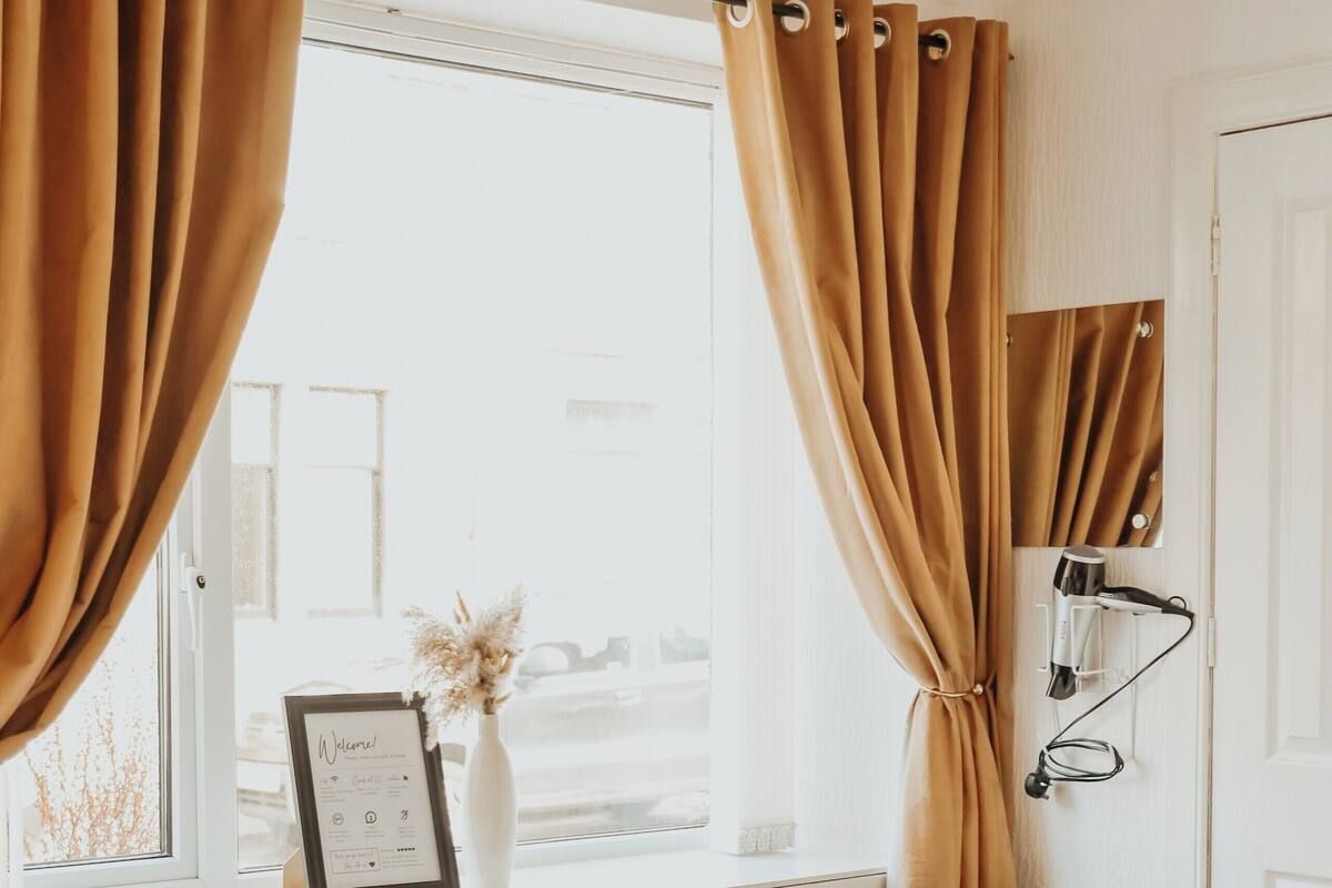 A hotel room with a bed, a chair, a window with curtains, a framed picture, and a vase with dried flowers.