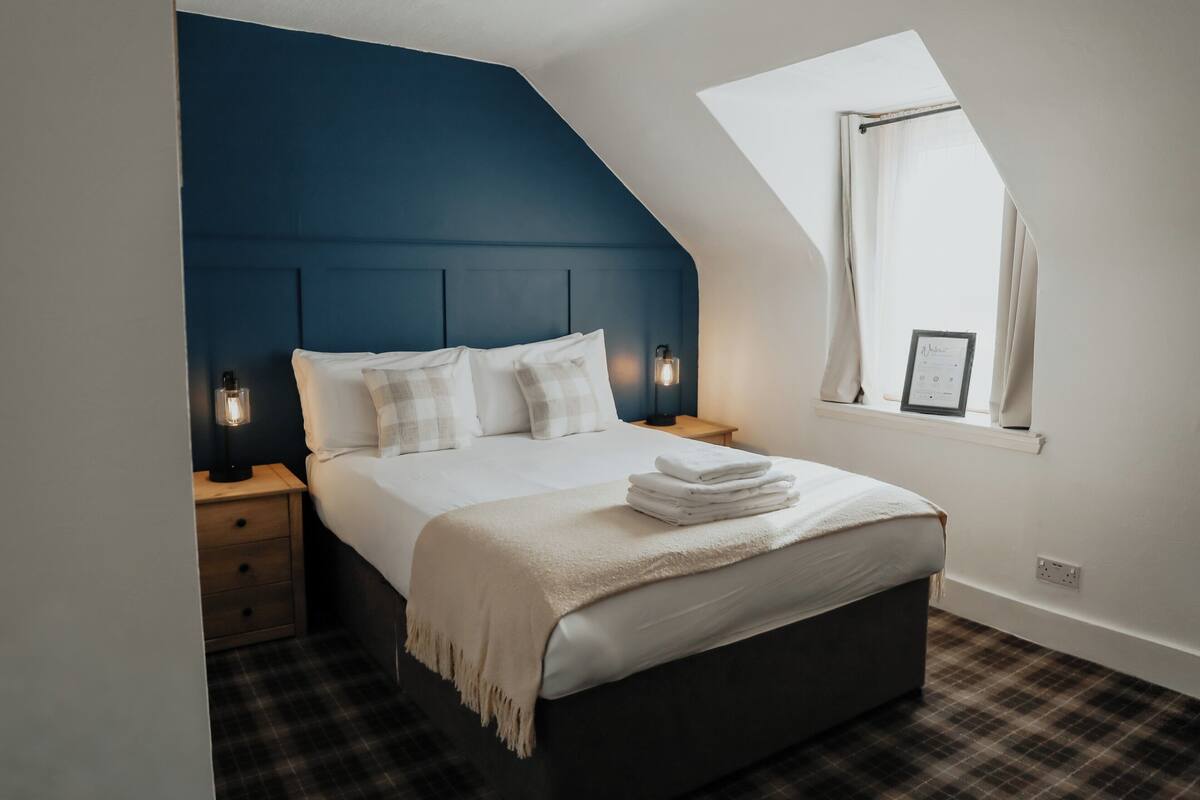 A bedroom with a dark blue headboard, a white bedspread, and a window with a view.