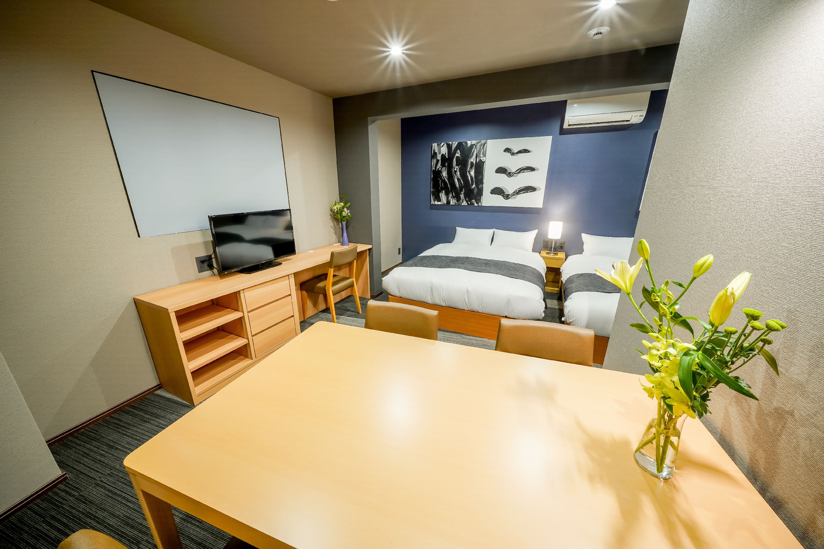 A hotel room with a bed, bedside table, TV, desk, and a vase of flowers.