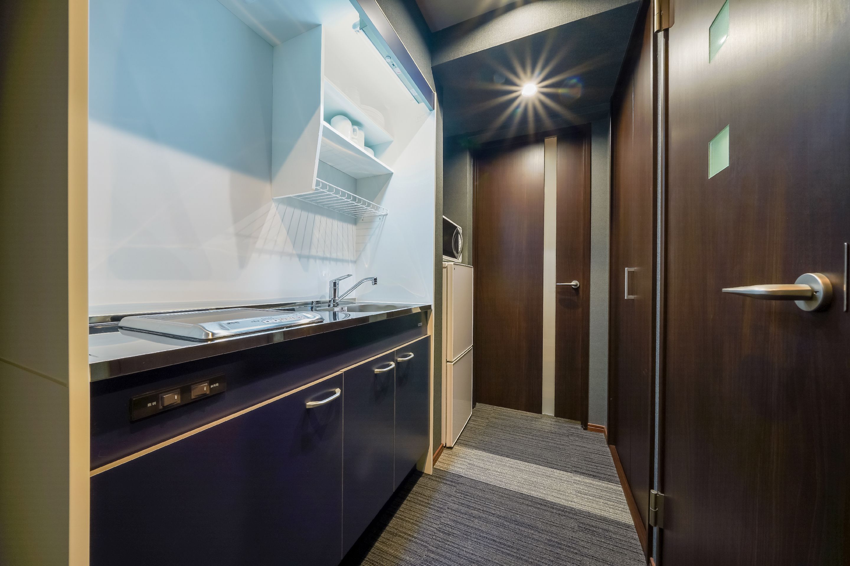 Una cocina moderna con horno empotrado, microondas y fregadero. Hay un refrigerador pequeño y un mueble con un estante sobre el fregadero. La cocina tiene un acabado de madera oscura y una alfombra a rayas que lleva a una puerta.