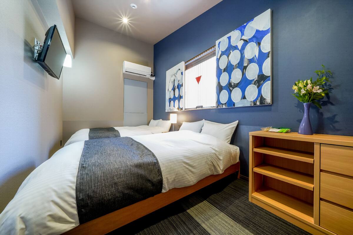 A hotel room with a bed, a desk, a chair, a wall-mounted TV, and artwork on the wall.