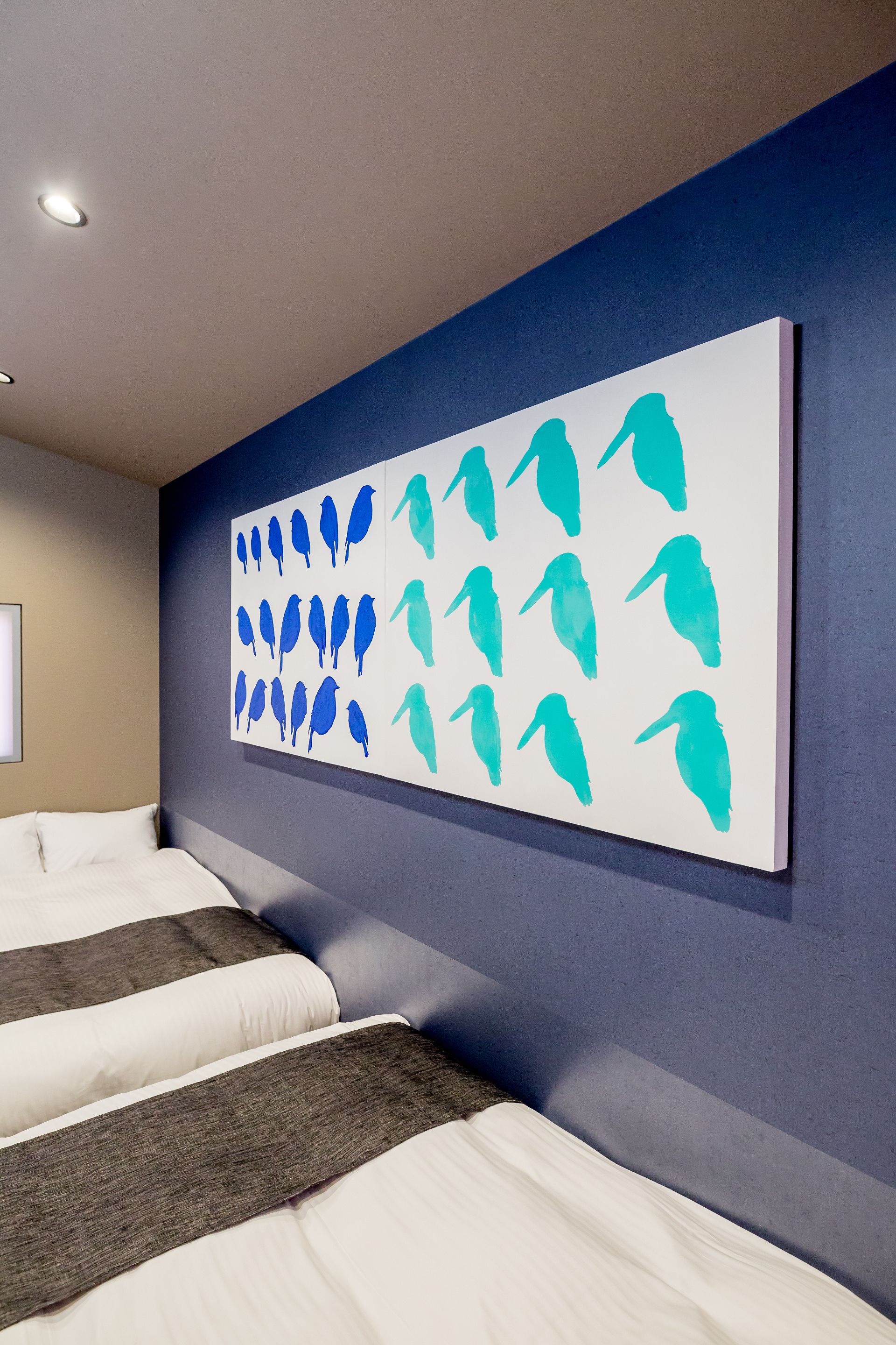 A modern hotel room with a large wall mural of stylized birds, two beds with white and gray bedding, and a dark blue accent wall.
