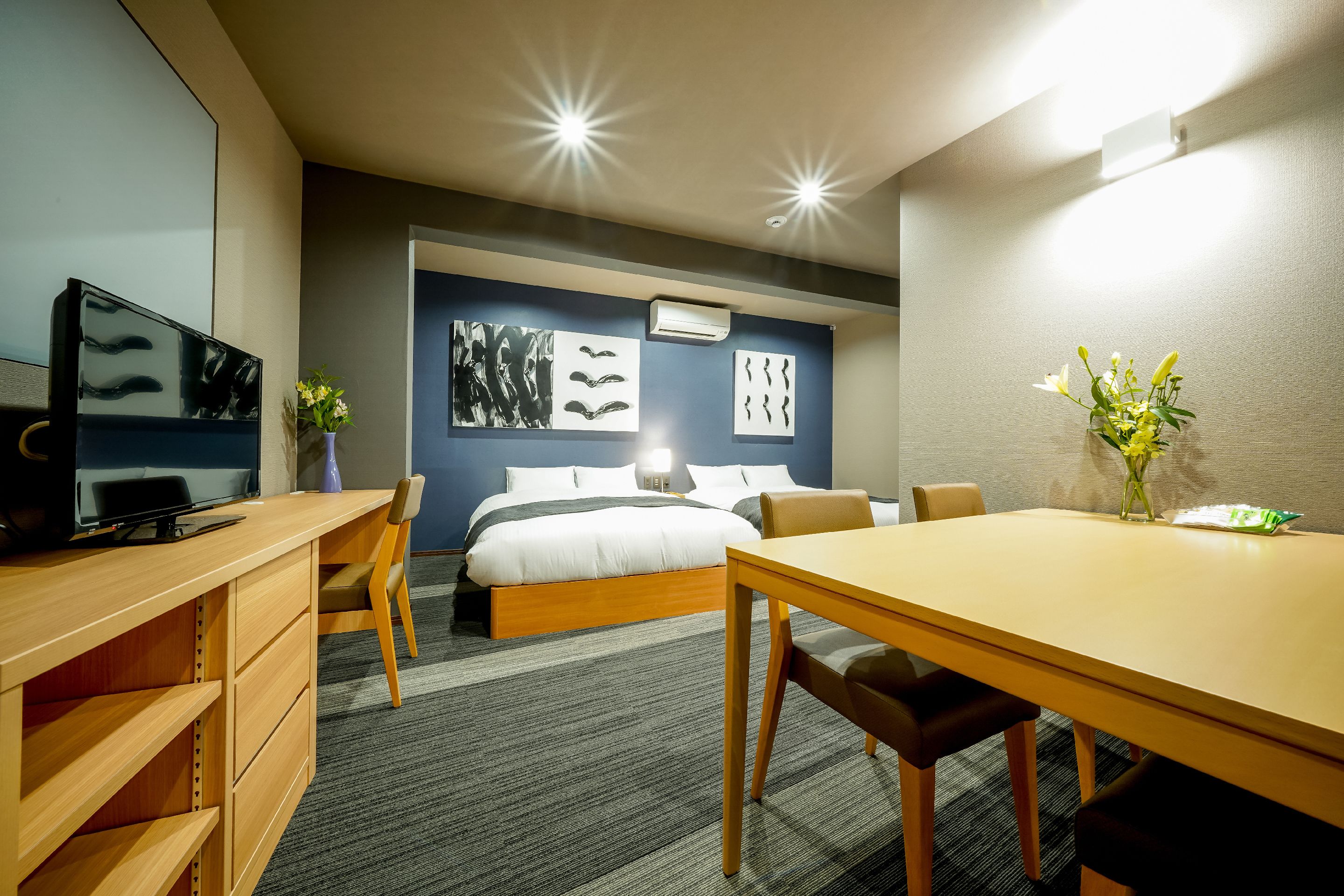 A hotel room with a bed, a desk with a TV, a dining table, and artwork on the walls.