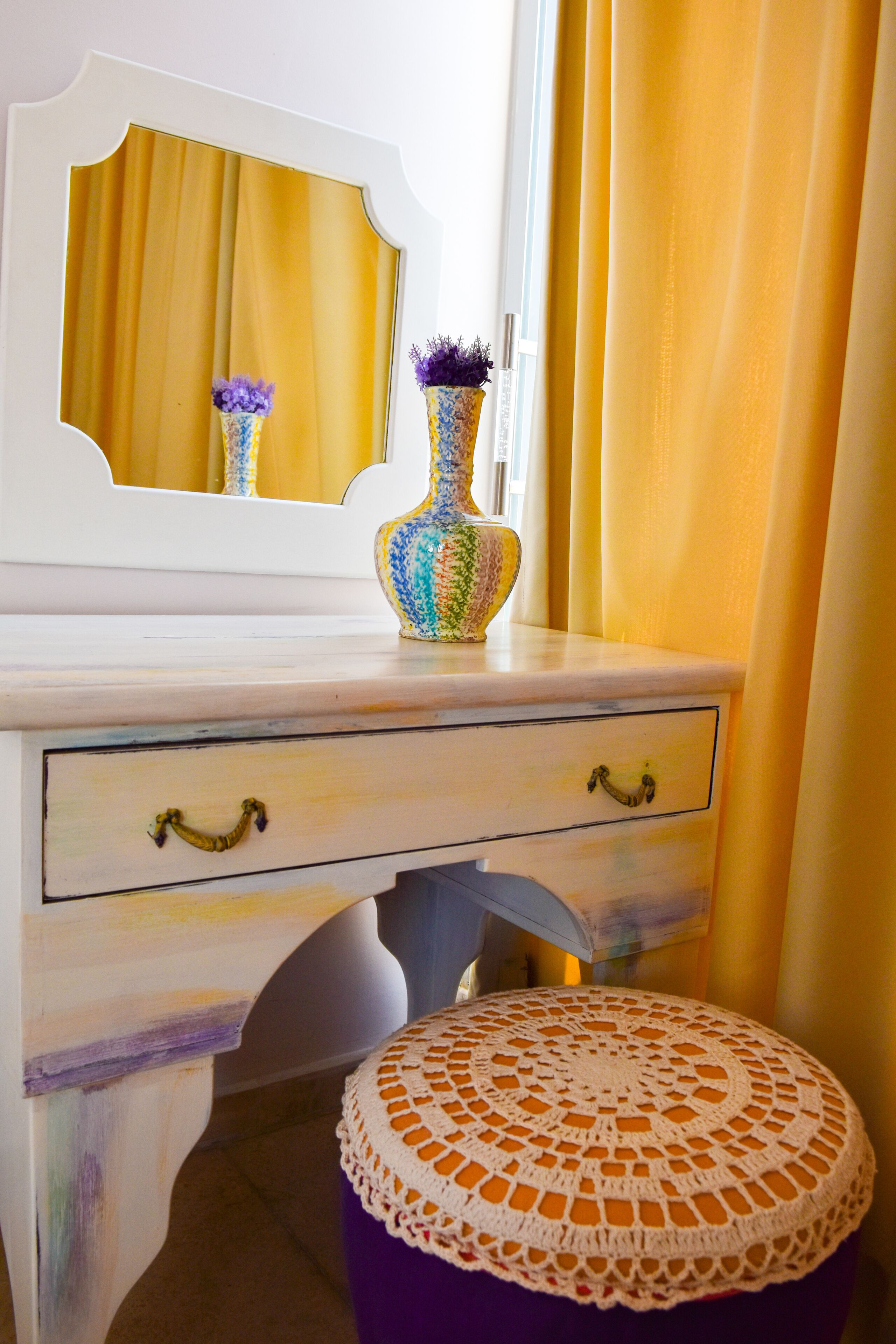 A white vanity with a mirror, a vase with purple flowers, and a purple stool with a crocheted cover.