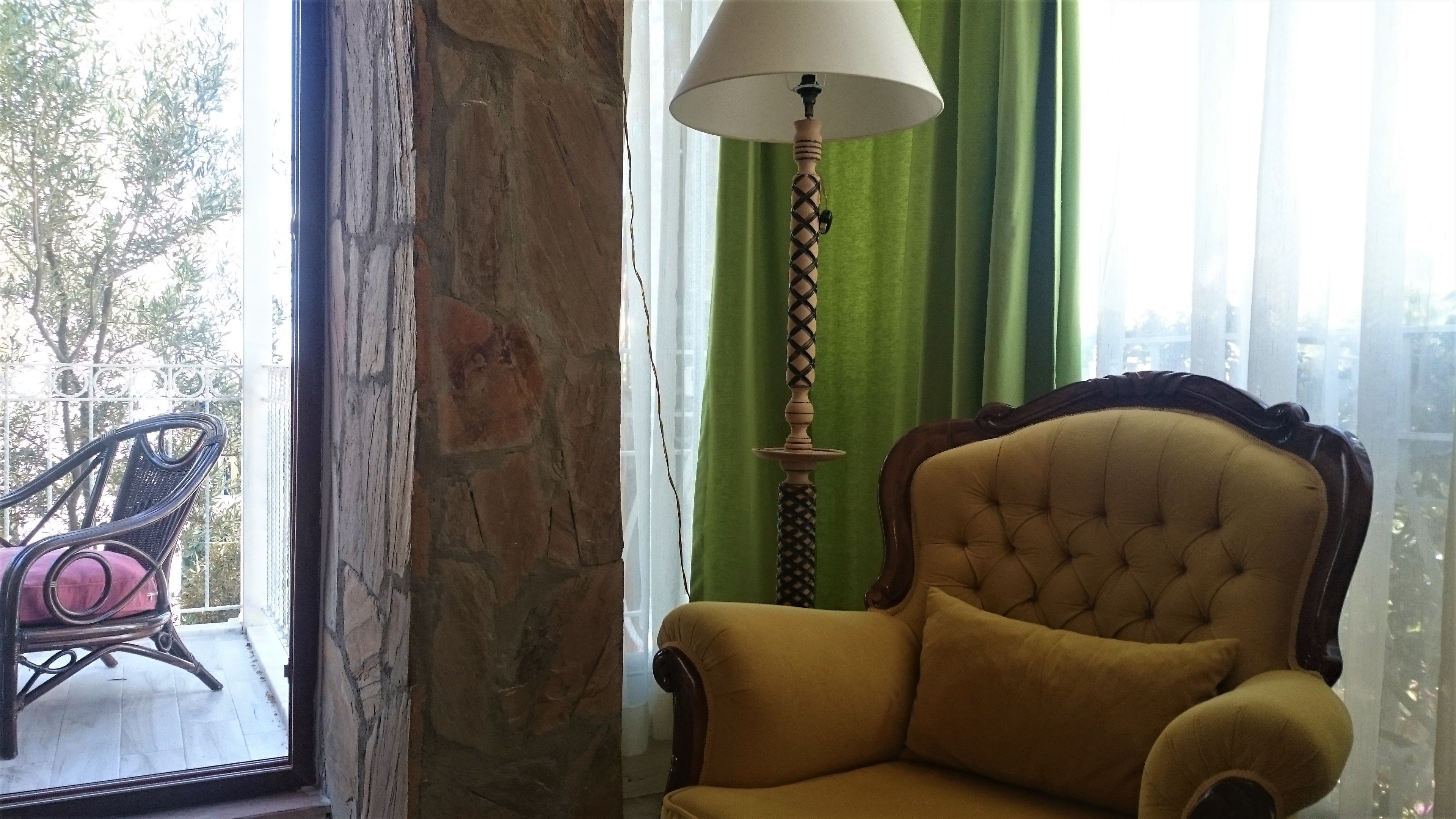 Une pièce avec un fauteuil jaune, un lampadaire et une vue sur l’extérieur par une fenêtre ornée de rideaux verts.