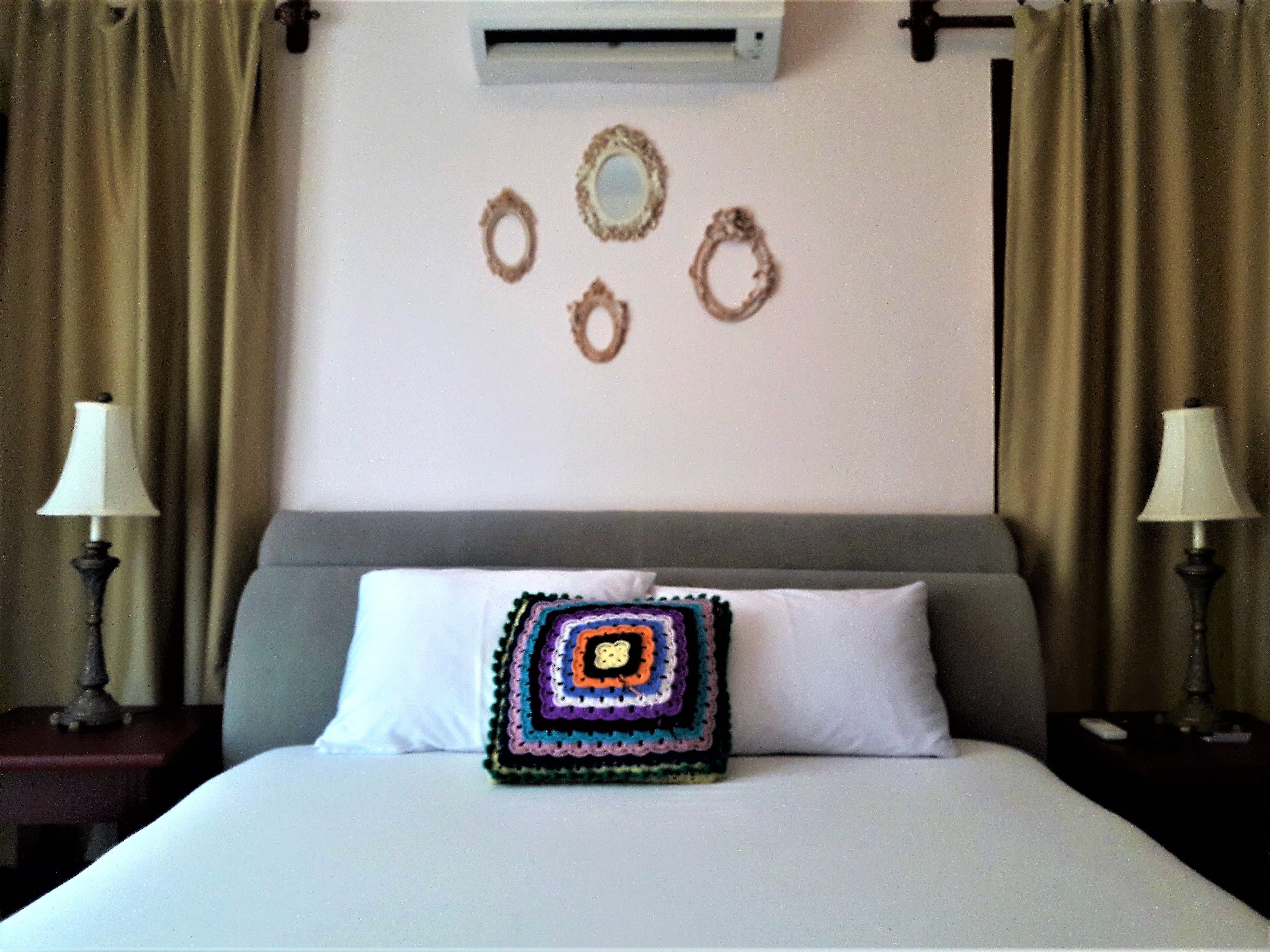 A hotel room with a bed, a decorative pillow, a wall air conditioner, and two lamps on nightstands.