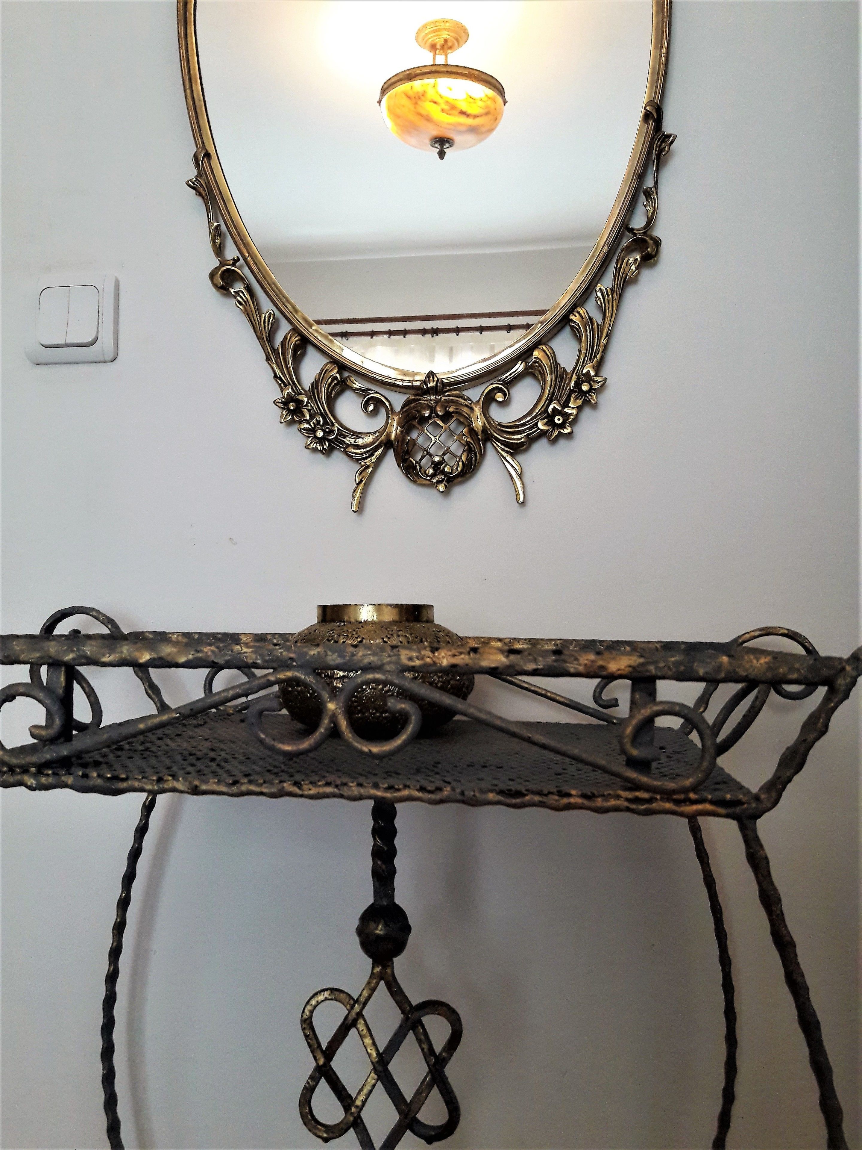 Ein Wandspiegel mit aufwendigem Rahmen und einer Leuchte darüber. Darunter ein Metalltisch mit dekorierter Tischplatte und einem hängenden Ornament.