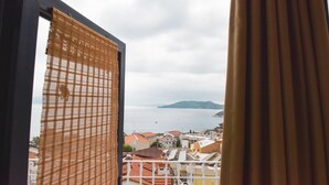 A view through a window with a bamboo blind, overlooking a coastal town with buildings and a body of water. - Smyrna Pansiyon (Kas)