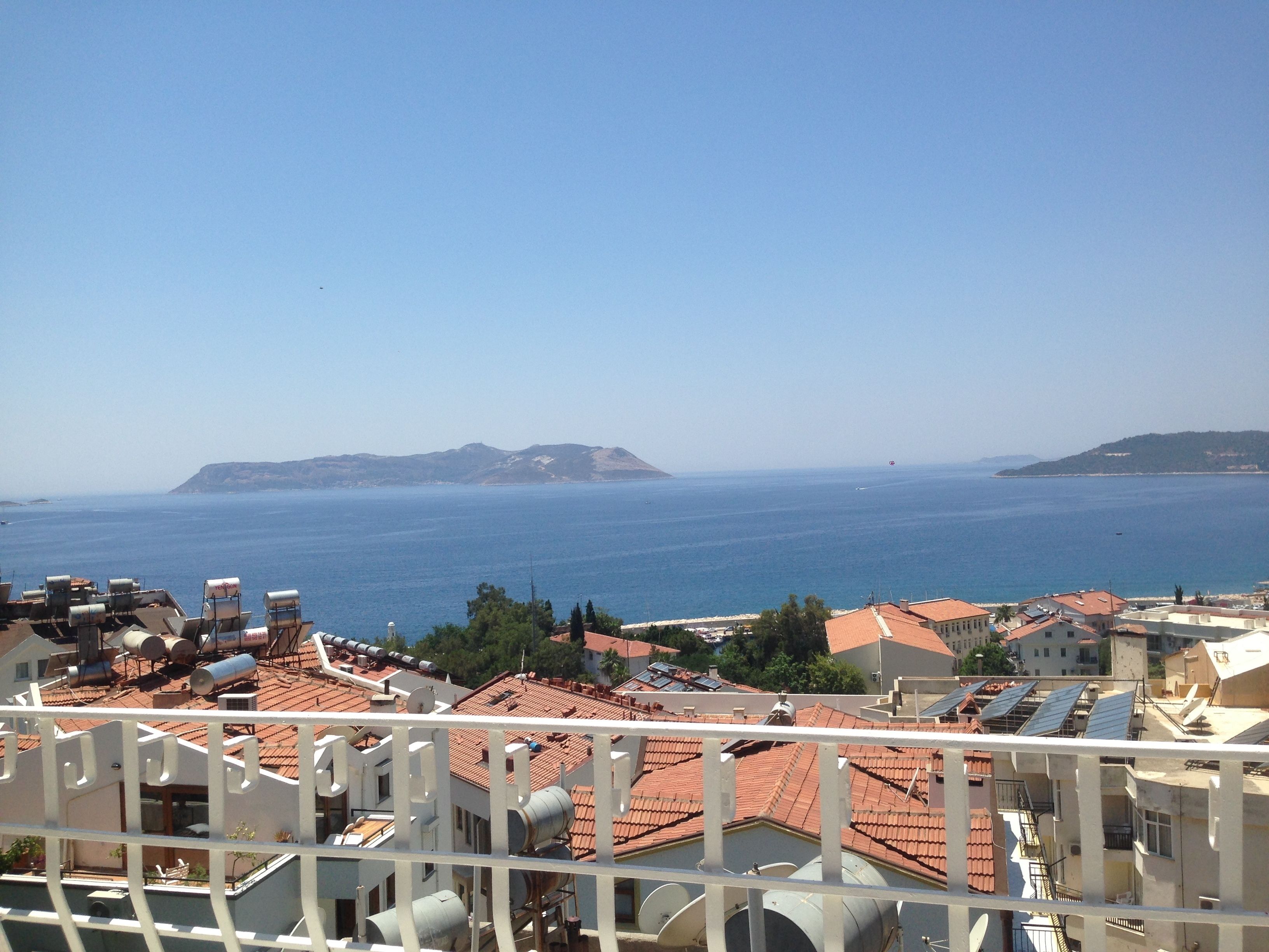 A coastal cityscape with terracotta-roofed buildings, a clear blue sea, and distant hills.