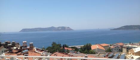 A coastal cityscape with terracotta-roofed buildings, a clear blue sea, and distant hills.