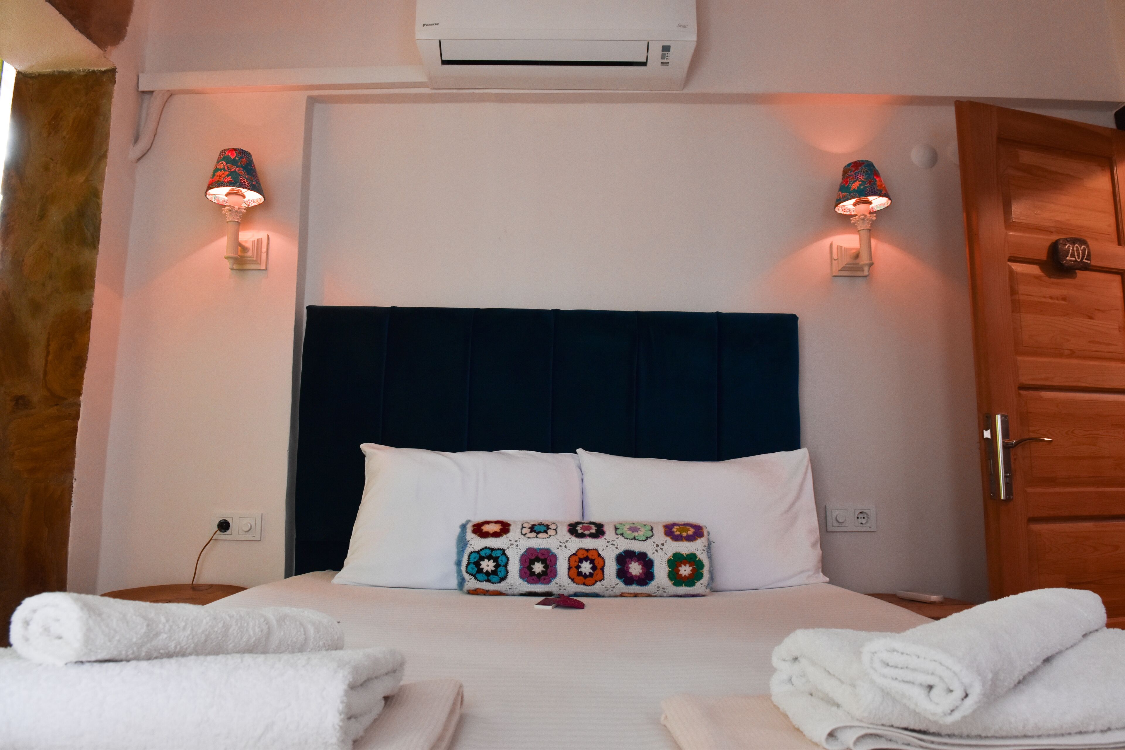 A hotel room with a bed, white towels, a decorative pillow, two wall-mounted lamps, and an air conditioning unit.