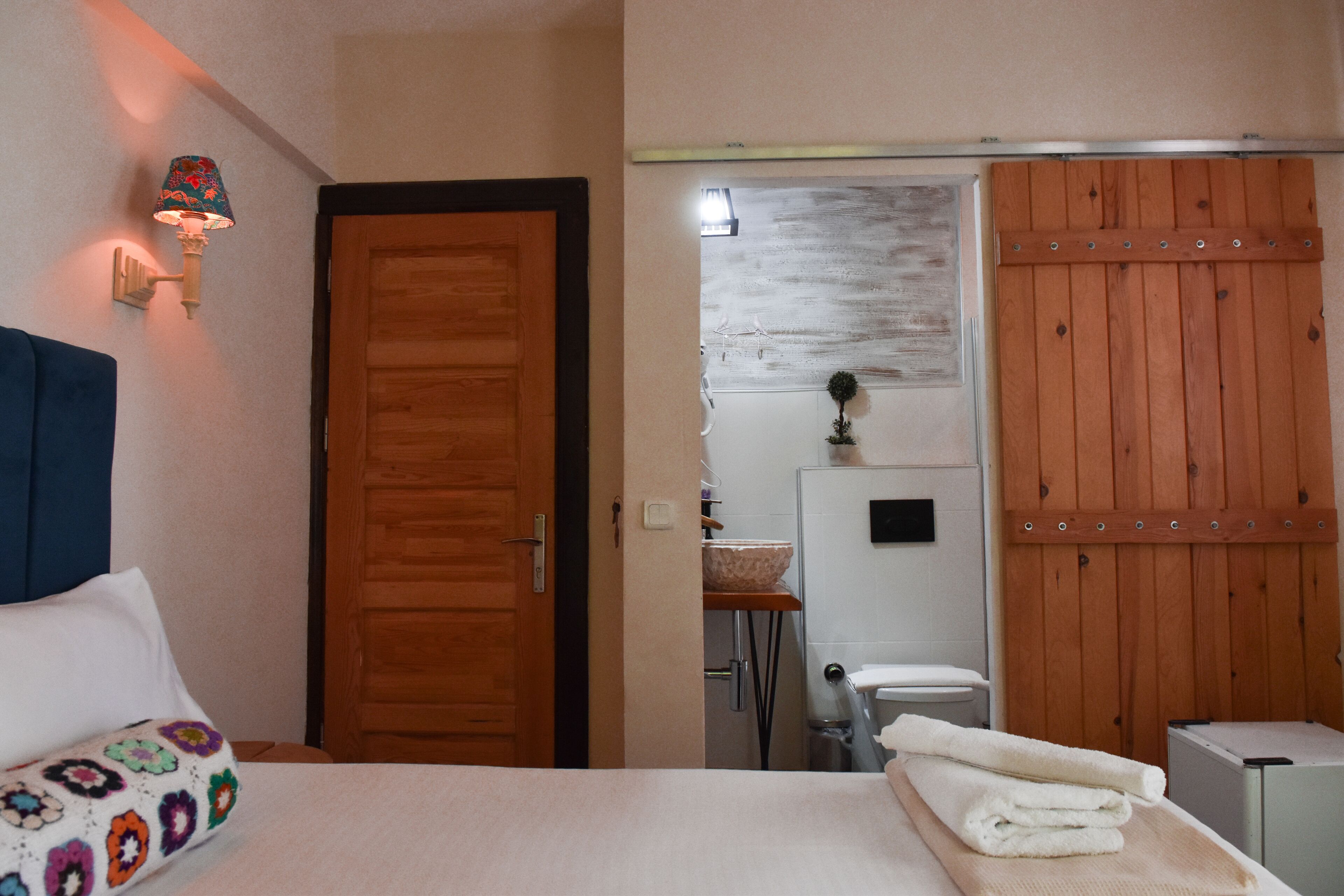 A hotel room with a wooden door, a bed with a colorful blanket, a bathroom with a wooden cabinet, and a towel on the floor.