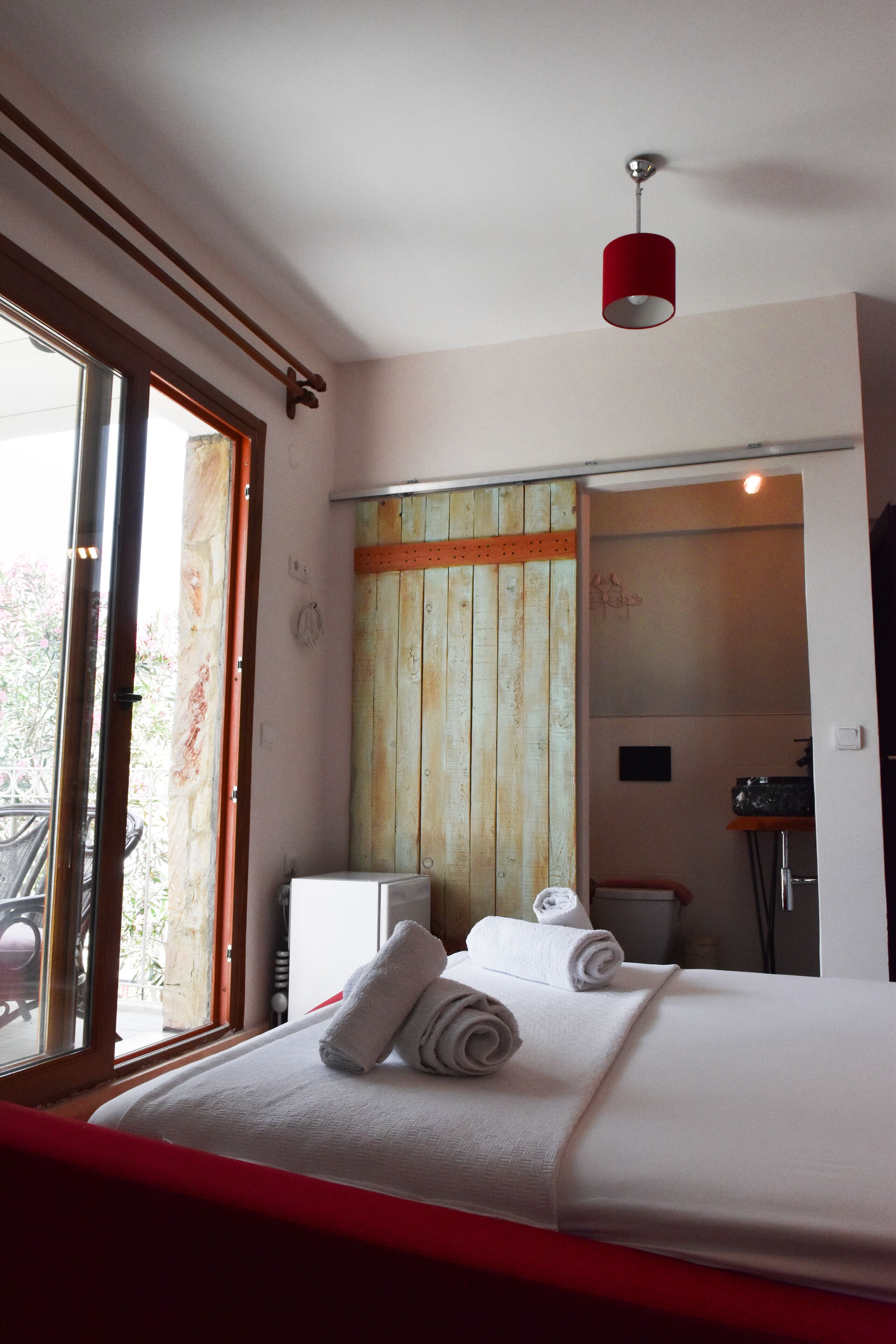 Una habitación de hotel con cama, ventana, una lámpara roja y pared de madera.