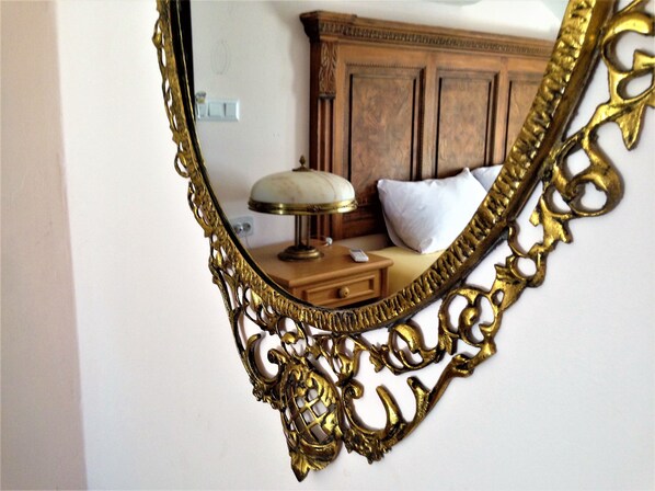 Un espejo con detalles ornamentales dorados refleja una habitación con un aparador de madera, una lámpara y una cama con almohadas.