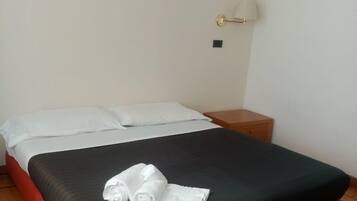 A neatly made bed with a black and red bedspread, white pillows, and a rolled white towel on the bed. There is a small bedside table with a lamp, a framed picture on the wall, and a wooden floor.