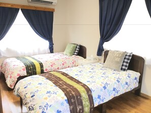 A bedroom with two single beds, each with floral bedding and a wooden headboard. There is a nightstand with a lamp and a tissue box on each bed. The room has wooden flooring and a window with blue curtains. - Pension Ishigaki Tomatamatsu (Ishigaki)