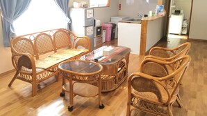 A room with a dining table surrounded by wicker chairs, a refrigerator, a microwave, and a small kitchen area. - Pension Ishigaki Tomatamatsu (Ishigaki)