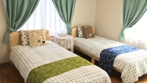 A room with two single beds, each with a patterned bedspread and pillows, a small round table with a tissue box, and a window with green curtains. - Pension Ishigaki Tomatamatsu (Ishigaki)