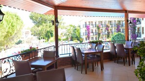 A covered outdoor seating area with tables and chairs, overlooking a courtyard with buildings and greenery.
