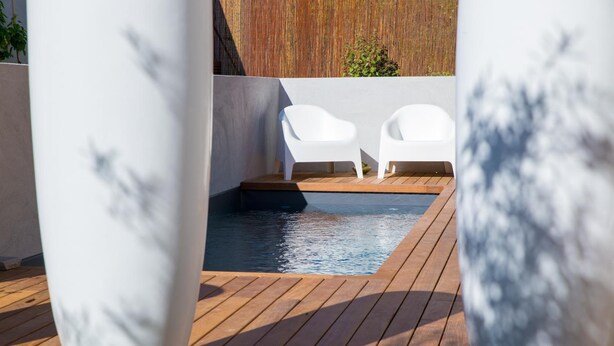 Una terraza de madera con una piscina pequeña, sillas blancas y una valla de bambú al fondo.