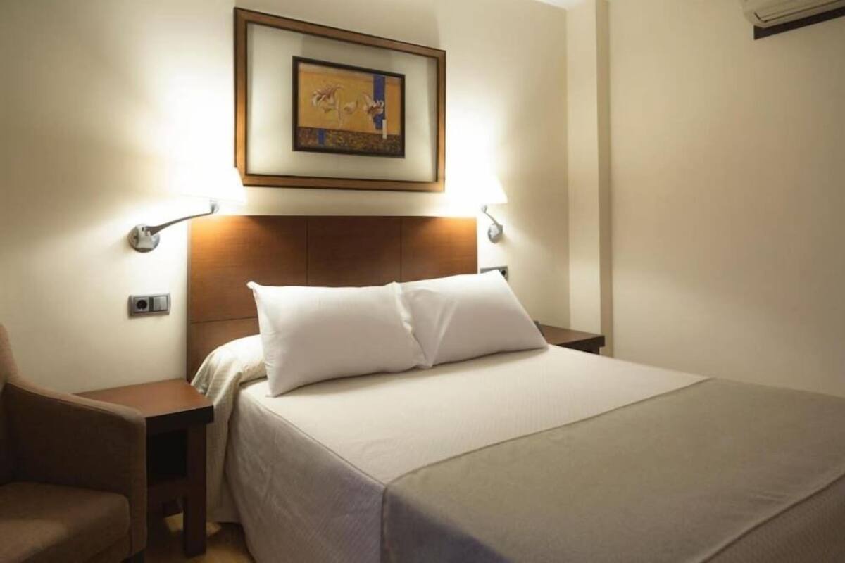 Quarto de hotel com cama, mesas de cabeceira, uma cadeira e um quadro na parede.