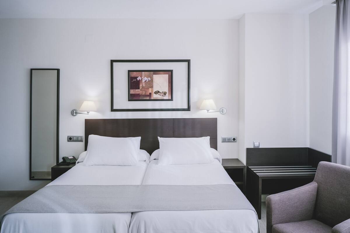 A hotel room with a double bed, bedside lamps, a nightstand, a chair, a framed picture on the wall, and a mirror.