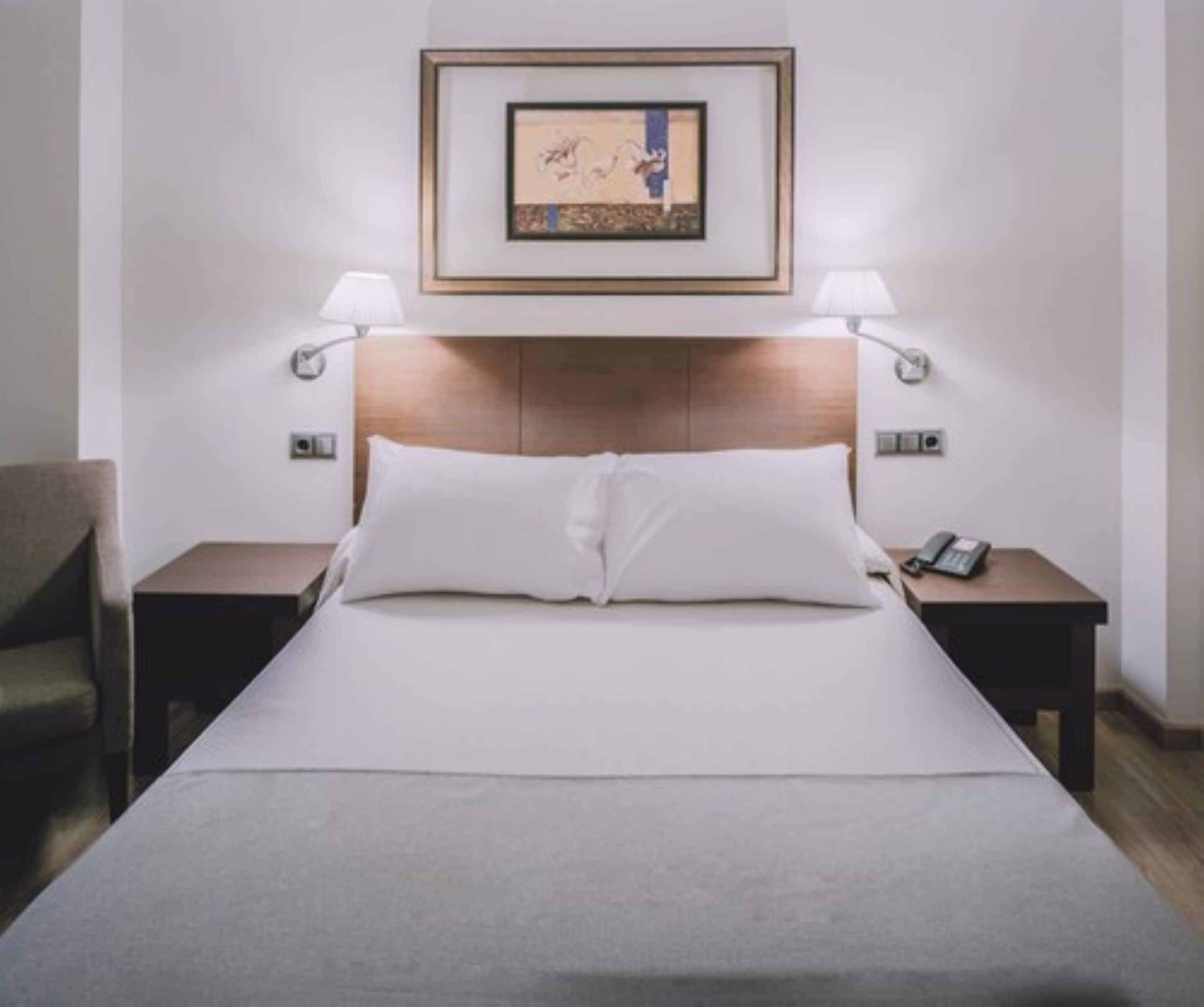 Una habitación de hotel con una cama, dos mesitas de noche, una silla, un cuadro enmarcado en la pared y una lámpara sobre cada mesita de noche.