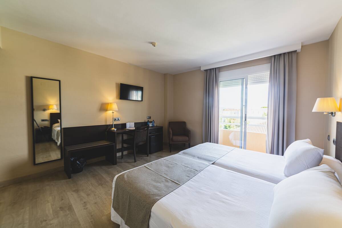 Quarto de hotel com cama, escrivaninha, cadeira e varanda com cortinas.