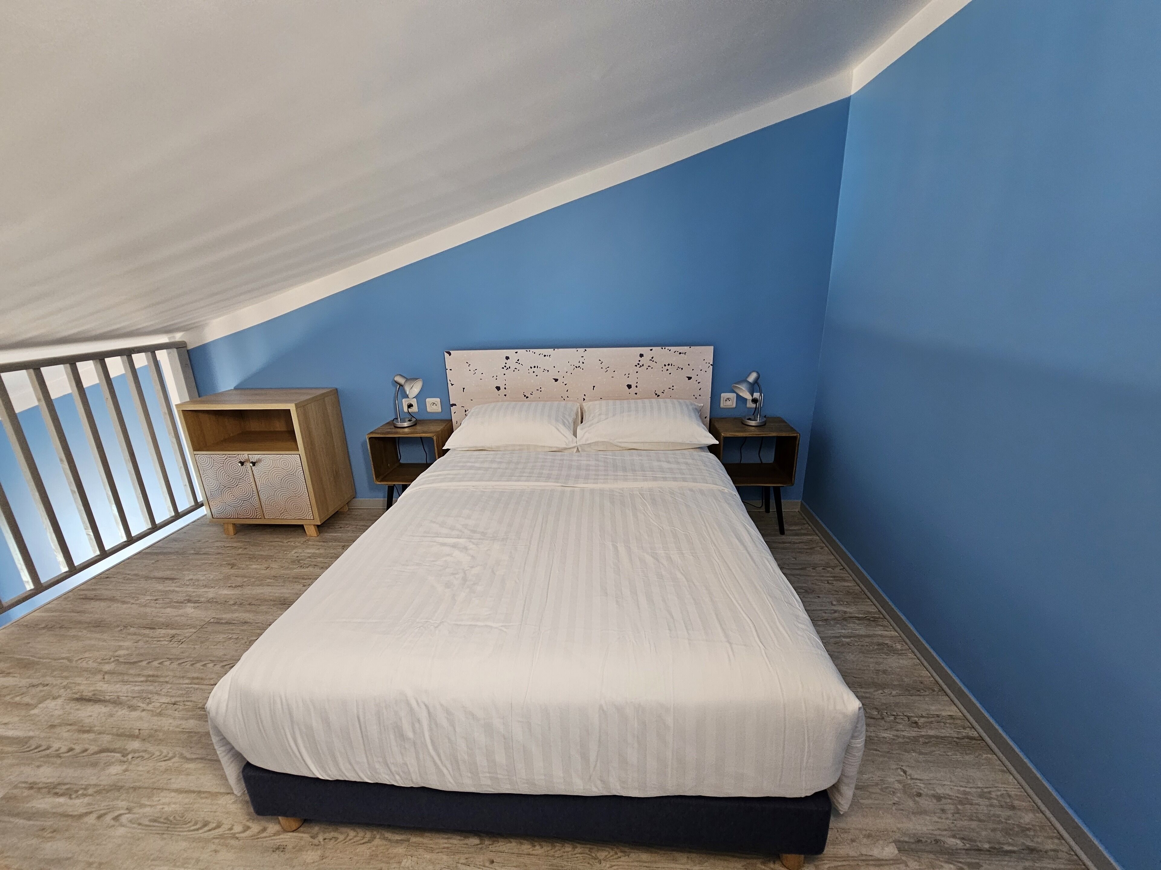 A bedroom with a bed, two bedside tables, a nightstand, a wardrobe, and a balcony with a railing.