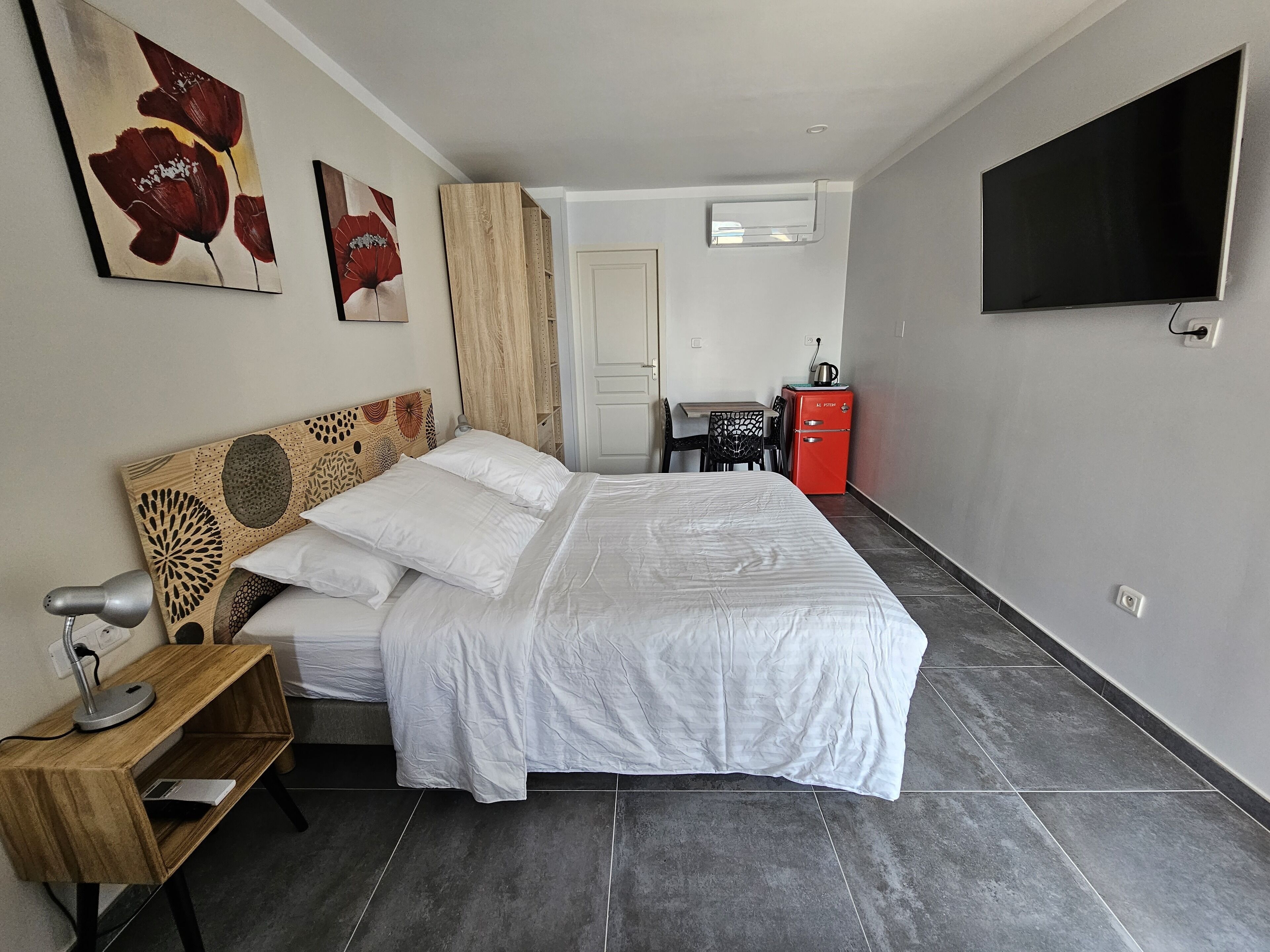 Un dormitorio ordenado con cama, escritorio, silla, televisión y una pequeña kitchenette.