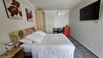 Un dormitorio ordenado con cama, escritorio, silla, televisión y una pequeña kitchenette.