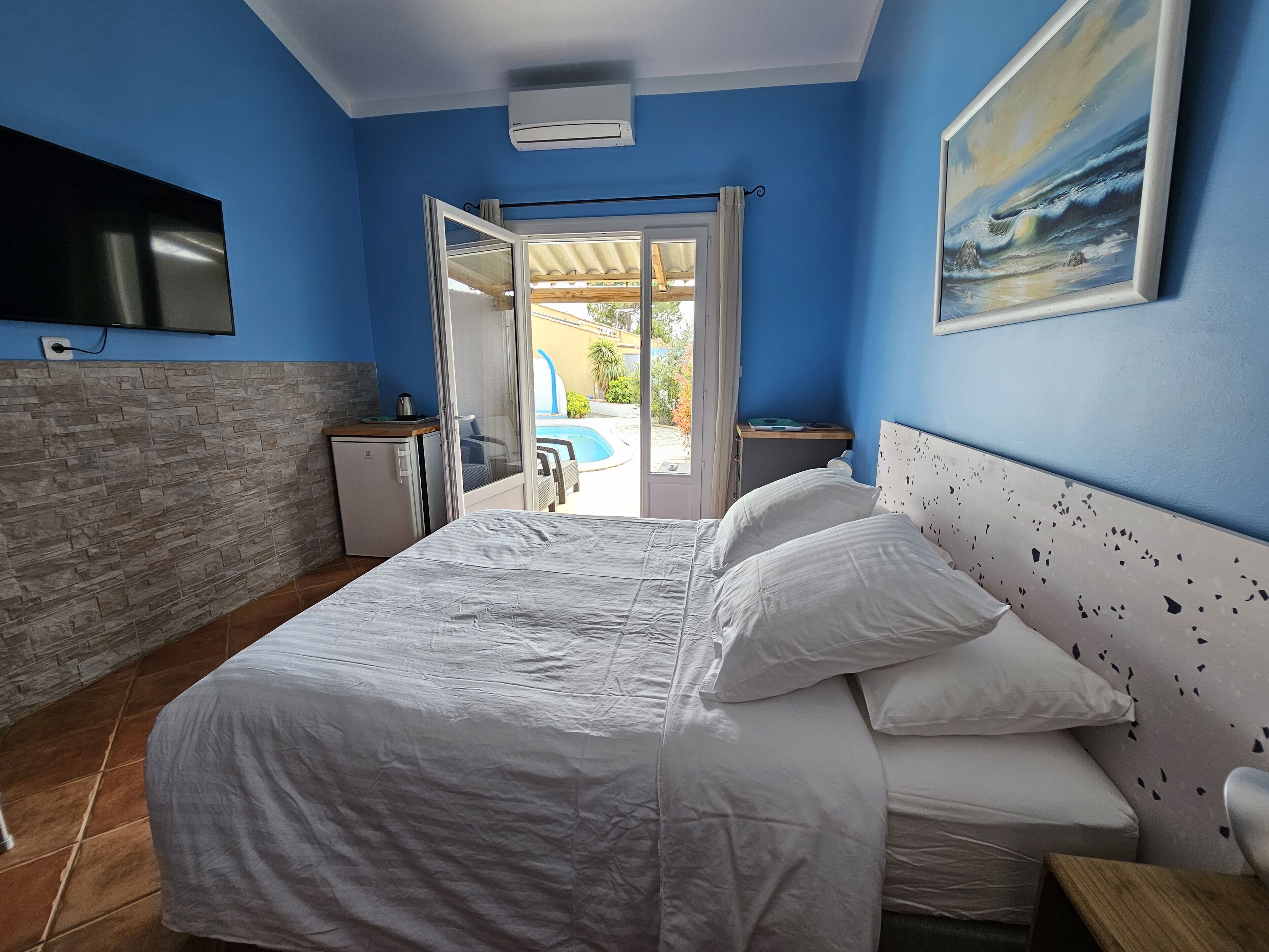 Een slaapkamer met een bed, een televisie, een kleine koelkast en een raam met uitzicht op een zwembad en terras.