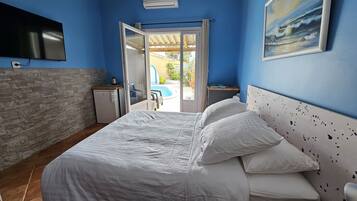 Een slaapkamer met een bed, een televisie, een kleine koelkast en een raam met uitzicht op een zwembad en terras.