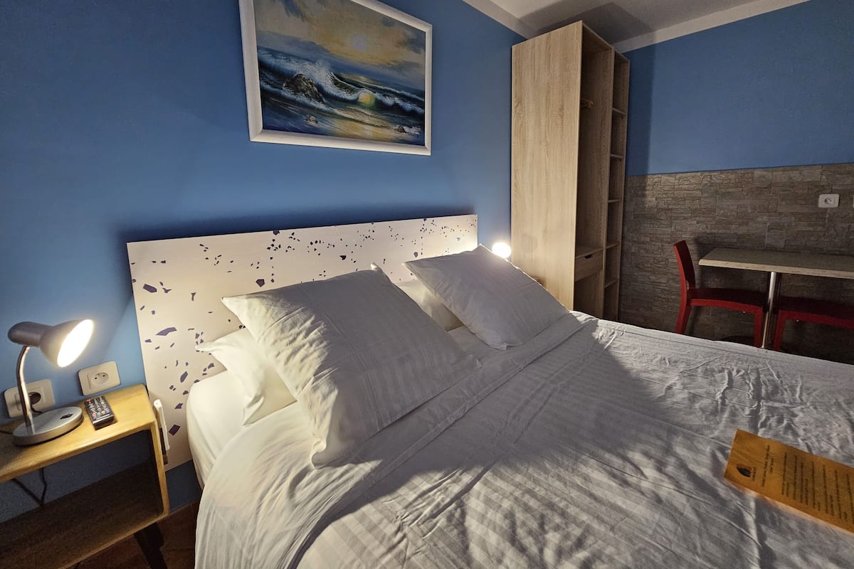 Een hotelkamer met een bed, een nachtkastje met een lamp, een schilderij aan de muur en een klein tafeltje met een stoel.