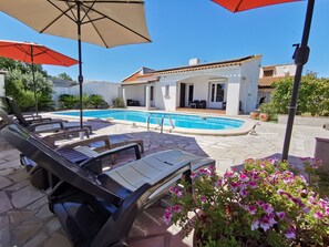 Un espace aménagé au bord de la piscine, avec des chaises longues, des parasols et un bâtiment doté d’un toit en tuiles.