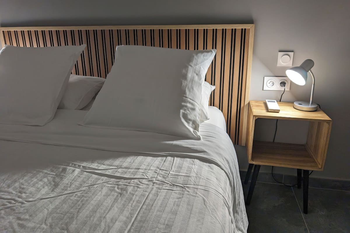 Een net opgemaakt bed met witte lakens en kussens, een houten hoofdbord met verticale latten, een nachtkastje met een lamp en een stopcontact.