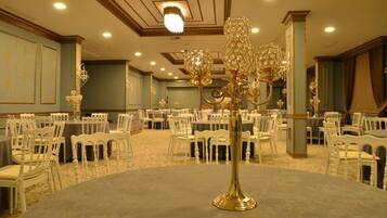 Uma sala de banquetes com mesas redondas, cadeiras brancas e lustres ornamentados.