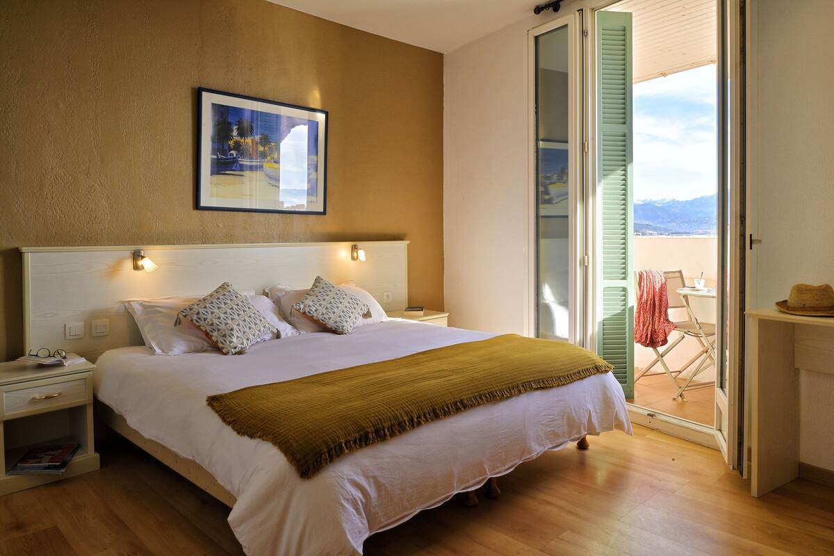 A bedroom with a large bed, a nightstand, a framed picture on the wall, and a balcony with a view.